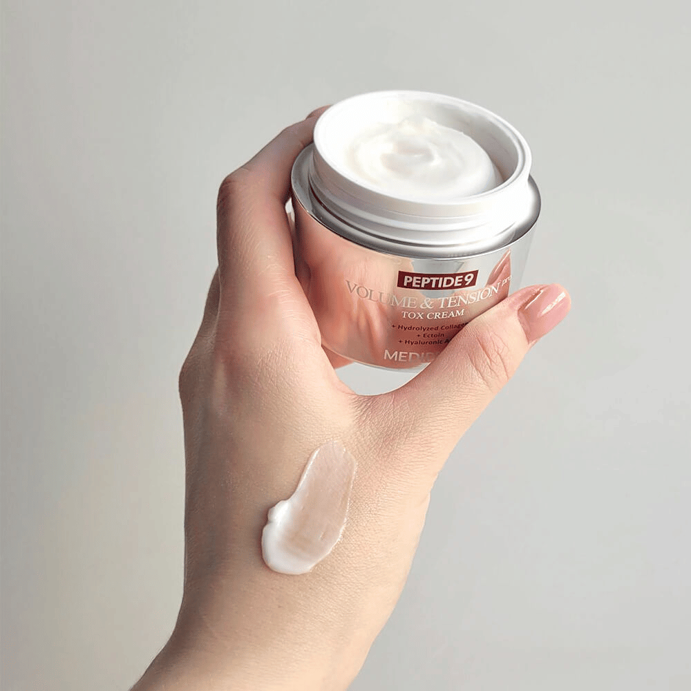 Χέρι κρατάει την MEDI-PEEL Peptide 9 Volume & Tension Tox Cream με μία σταγόνα κρέμας στο πίσω μέρος του χεριού. Η μεταξένια φόρμουλα ενυδατώνει και προσφέρει στο δέρμα σφριγηλότητα και ελαστικότητα.