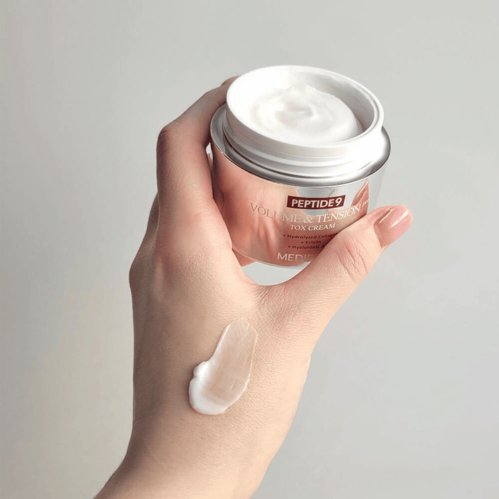 Hand håller i MEDI-PEEL Peptide 9 Volume & Tension Tox Cream med en klick kräm på handryggen. Den silkeslena formulan återfuktar och ger huden fasthet och spänst.