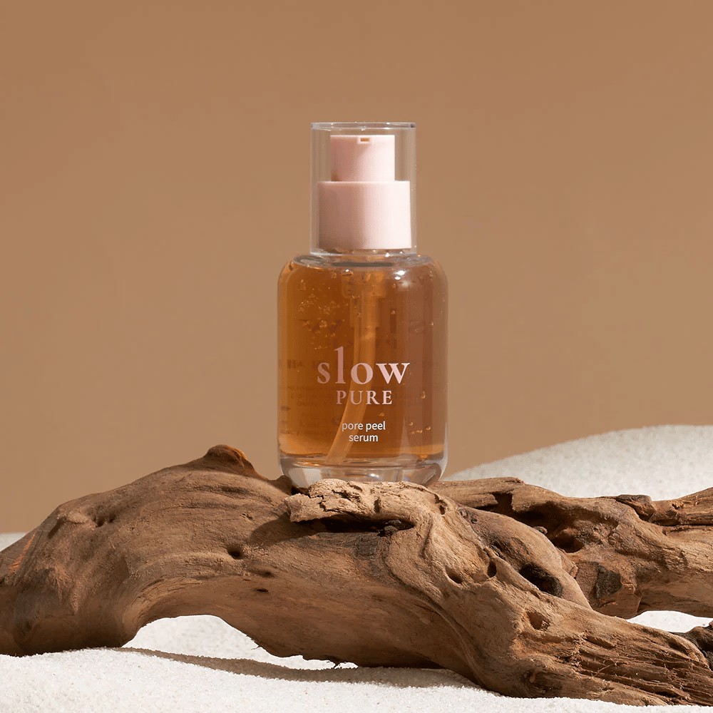 Slow Pure Pore Peel Serum τοποθετημένο στην άμμο με ξύλινη λεπτομέρεια στο φόντο, σύμβολο φυσικής περιποίησης δέρματος.