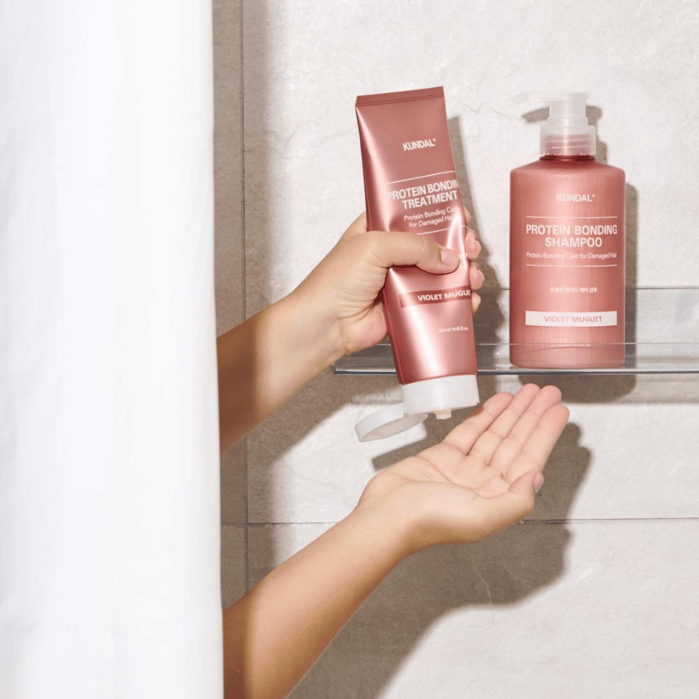 KUNDAL Protein Bonding Treatment Violet Muguet χρησιμοποιείται στο ντους, με το Protein Bonding Shampoo τοποθετημένο στο ράφι δίπλα.