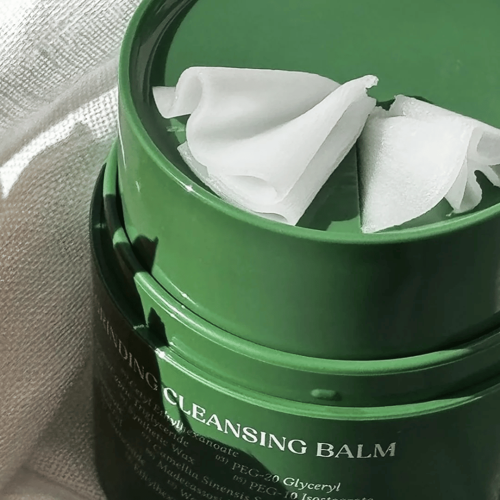 Η εικόνα δείχνει ένα πράσινο δοχείο με "Cleansing Balm", το οποίο είναι ανοιχτό για να αποκαλύψει τη συνοδευτική σπάτουλα και το απαλό περιεχόμενο. Η κρεμώδης υφή αντανακλά το φως, προσφέροντας μια αίσθηση πολυτέλειας και αποτελεσματικότητας στον καθαρισμό.