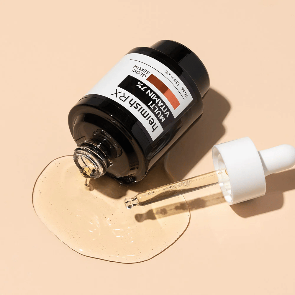 Heimish RX Πολυβιταμίνη 7% Glow Serum ξαπλωμένη σε μπεζ φόντο με τον ορό να είναι απλωμένος γύρω από το μπουκάλι και μια πιπέτα γεμάτη με προϊόν, που αναδεικνύει τη θρεπτική του φόρμουλα.