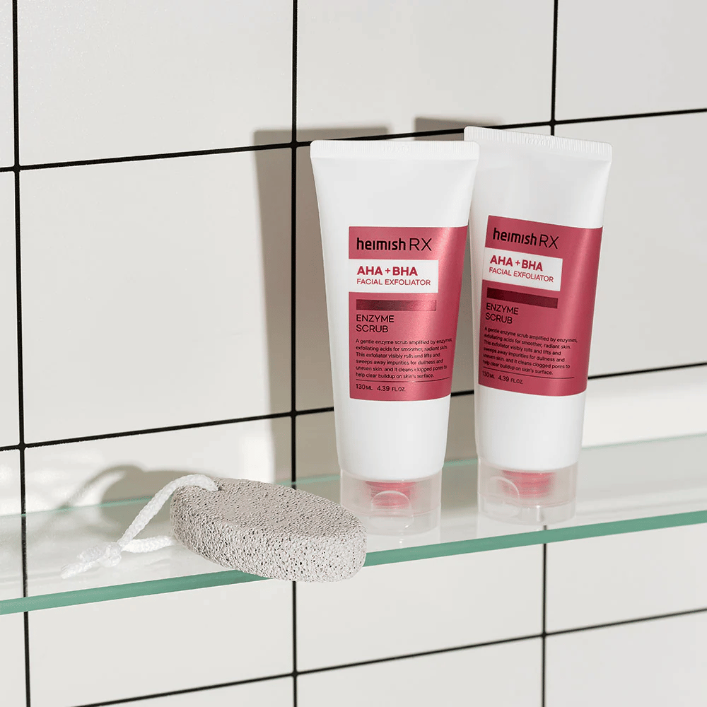 Δύο σωλήνες Heimish RX AHA + BHA Facial Exfoliator τοποθετημένοι σε γυάλινο ράφι σε μπάνιο με λευκό τοίχο από πλακάκια. Δίπλα στα προϊόντα υπάρχει μια ελαφρόπετρα με λευκό κορδόνι. Μια κομψή και μινιμαλιστική εικόνα περιποίησης δέρματος.