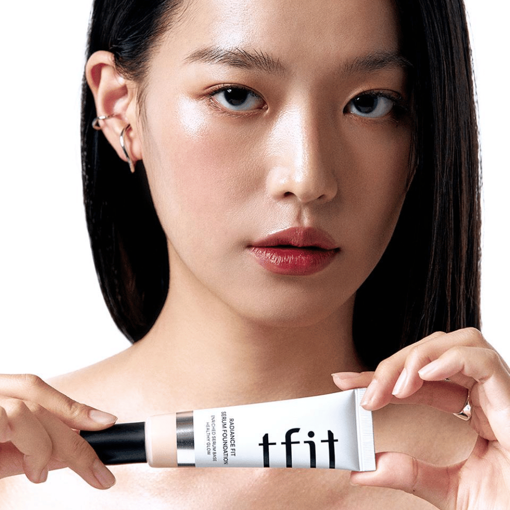 Μοντέλο με γυαλιστερό, λείο δέρμα κρατάει TFIT Radiance Fit Serum Foundation, το οποίο αναδεικνύει το φυσικό φινίρισμα και τη λάμψη του προϊόντος.