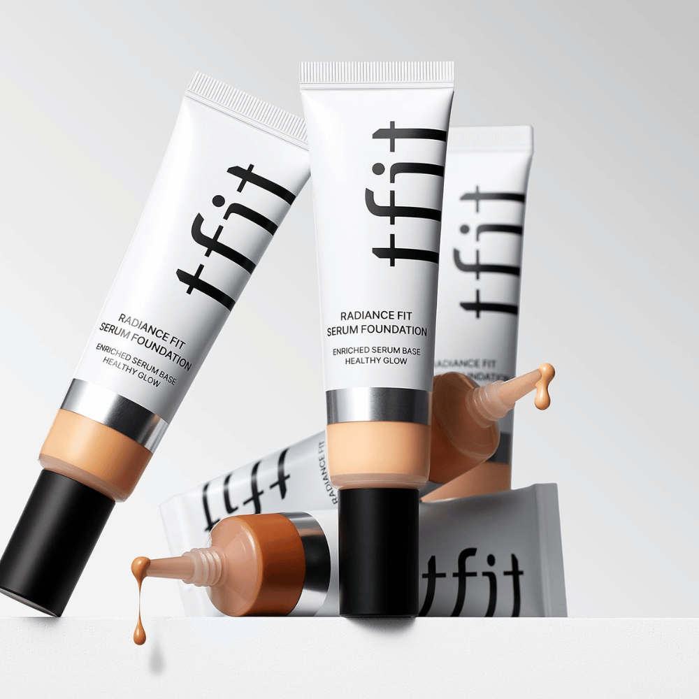 Πολλοί σωλήνες από TFIT Radiance Fit Serum Foundation σε διάφορες αποχρώσεις, εκ των οποίων ένας με foundation που στάζει, σε ανοιχτό γκρι φόντο.