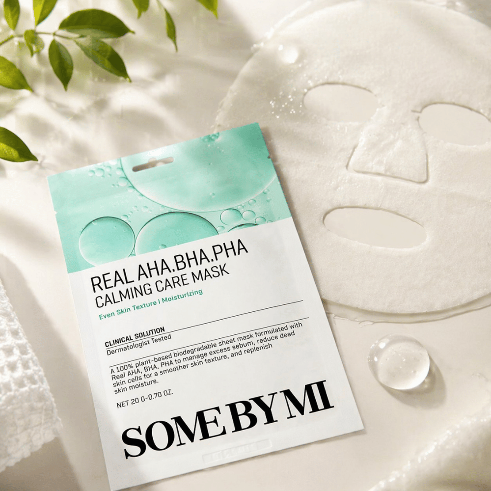 Η SOME BY MI Real AHA.BHA.PHA Calming Care Mask παρουσιάζεται μαζί με την ανεπτυγμένη υφασμάτινη μάσκα. Γύρω από το προϊόν φαίνονται σταγόνες υγρασίας και πράσινα φύλλα που ενισχύουν την αίσθηση ενυδάτωσης, καταπραϋντικής φροντίδας και ήπιας απολέπισης. Η εικόνα αναδεικνύει την έμφαση της μάσκας σε πιο ομοιόμορφη υφή της επιδερμίδας και βελτιωμένη ισορροπία υγρασίας.