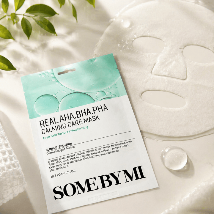Η SOME BY MI Real AHA.BHA.PHA Calming Care Mask παρουσιάζεται μαζί με την ανεπτυγμένη υφασμάτινη μάσκα. Γύρω από το προϊόν φαίνονται σταγόνες υγρασίας και πράσινα φύλλα που ενισχύουν την αίσθηση ενυδάτωσης, καταπραϋντικής φροντίδας και ήπιας απολέπισης. Η εικόνα αναδεικνύει την έμφαση της μάσκας σε πιο ομοιόμορφη υφή της επιδερμίδας και βελτιωμένη ισορροπία υγρασίας.