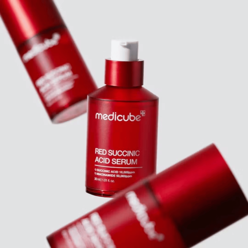 Medicube Red Succinic Acid Serum σε κόκκινο μπουκάλι με αντλία, περιτριγυρισμένο από περισσότερα όμοια μπουκάλια σε φωτεινό φόντο. Το προϊόν περιέχει σουκινικό οξύ και νιασιναμίδη για τη θεραπεία της ακμής και των χρωματικών κηλίδων.