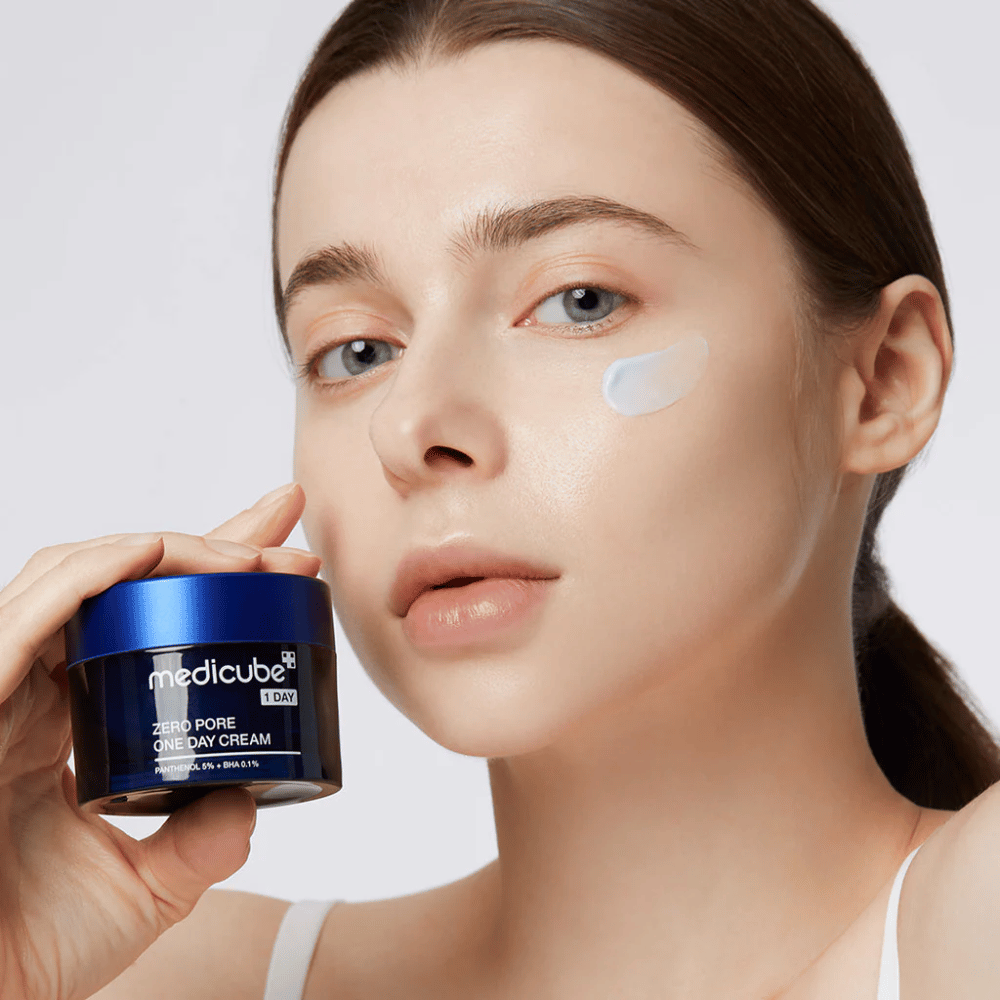 Γυναίκα με φυσική εμφάνιση κρατάει Medicube Zero Pore One Day Cream. Μια μικρή ποσότητα κρέμας είναι εφαρμοσμένη στο μάγουλό της. Το προϊόν έχει σχεδιαστεί για να βελτιώσει την υφή του δέρματος και να ελαχιστοποιήσει το μέγεθος των πόρων, με μια ελαφριά και ενυδατική φόρμουλα.