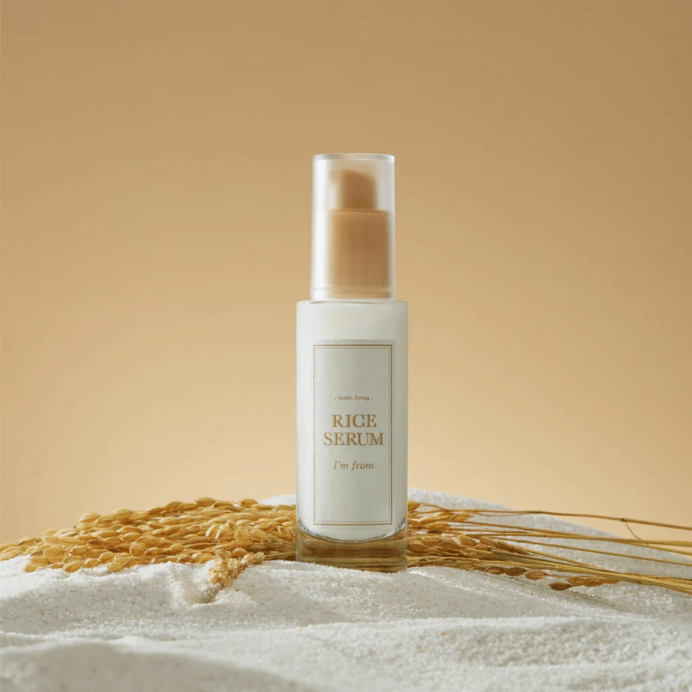 Μπουκάλι με "I'm from" Rice Serum, περιτριγυρισμένο από κόκκους ρυζιού και στάχυα ρυζιού σε ζεστό φόντο.