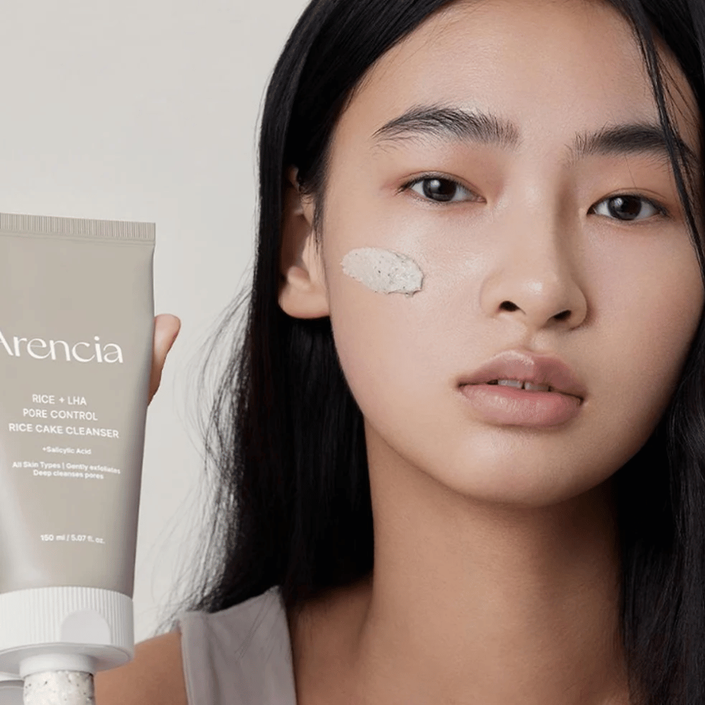 Μοντέλο που κρατάει Arencia Rice + LHA Pore Control Rice Cake Cleanser με εφαρμοσμένο προϊόν στο μάγουλο, απεικονίζει ήπιο αλλά αποτελεσματικό καθαρισμό πόρων για καθημερινή χρήση.