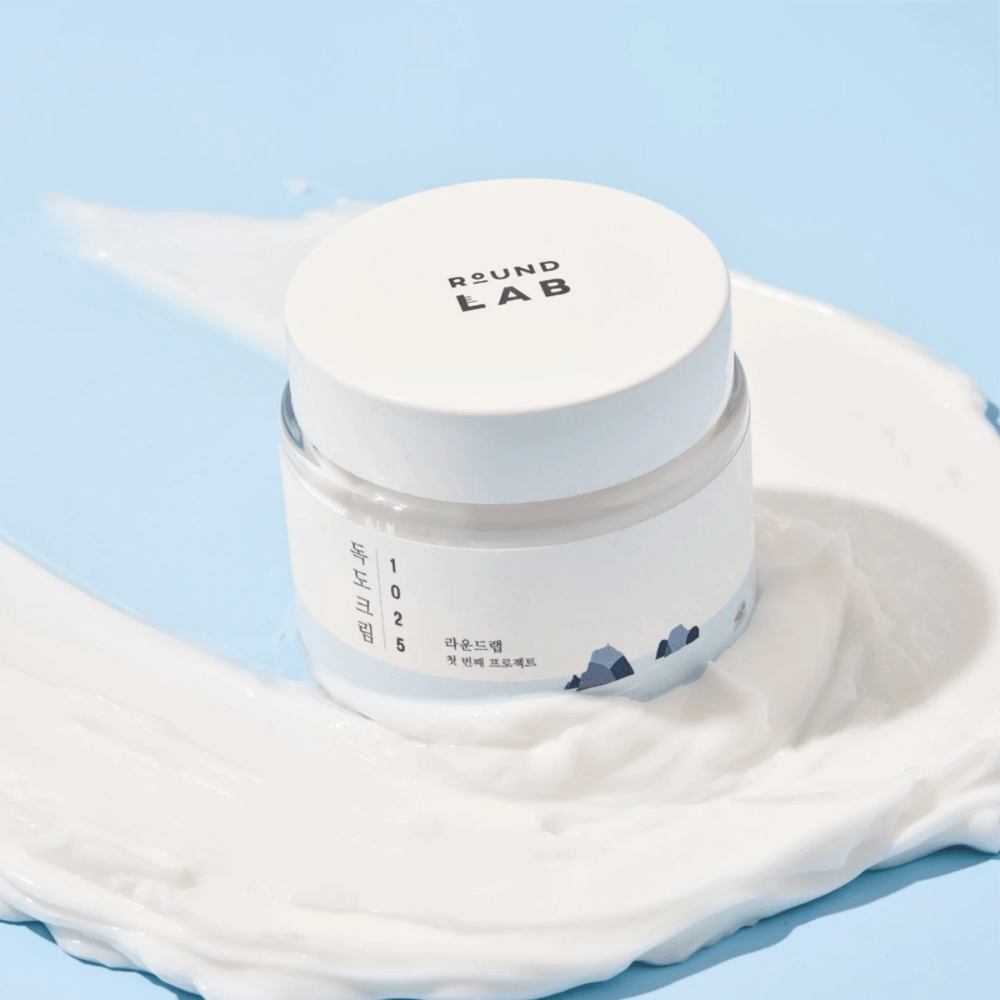 Ένα βάζο με Round Labs 1025 Dokdo Cream 80 ml που παρέχει ενυδάτωση, τοποθετημένο σε ζωντανό μπλε φόντο.