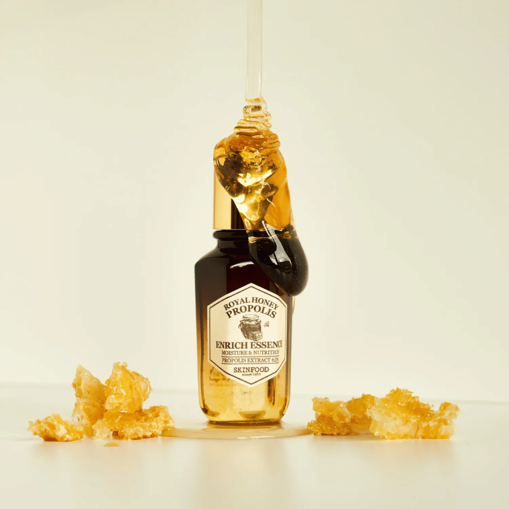 Εικόνα ενός γυάλινου μπουκαλιού με Royal Honey Propolis Enrich Essence από Skinfood. Μια μεγάλη σταγόνα χρυσού μελιού κυλάει από το χρυσό καπάκι του μπουκαλιού. Γύρω από το μπουκάλι βρίσκονται μικρά κομμάτια κηρήθρας σκορπισμένα σε μια φωτεινή επιφάνεια. Η ουσία προωθείται με έμφαση στην υγρασία και τη διατροφή, με 63% εκχύλισμα πρόπολης.