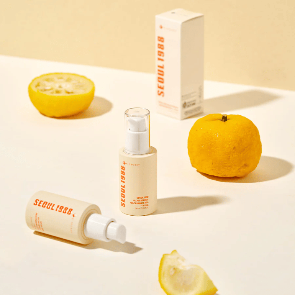 Πολλά μπουκάλια και συσκευασίες του Seoul 1988 Glow Serum τοποθετημένα με ολόκληρα και μισά φρούτα yuja στο παρασκήνιο.