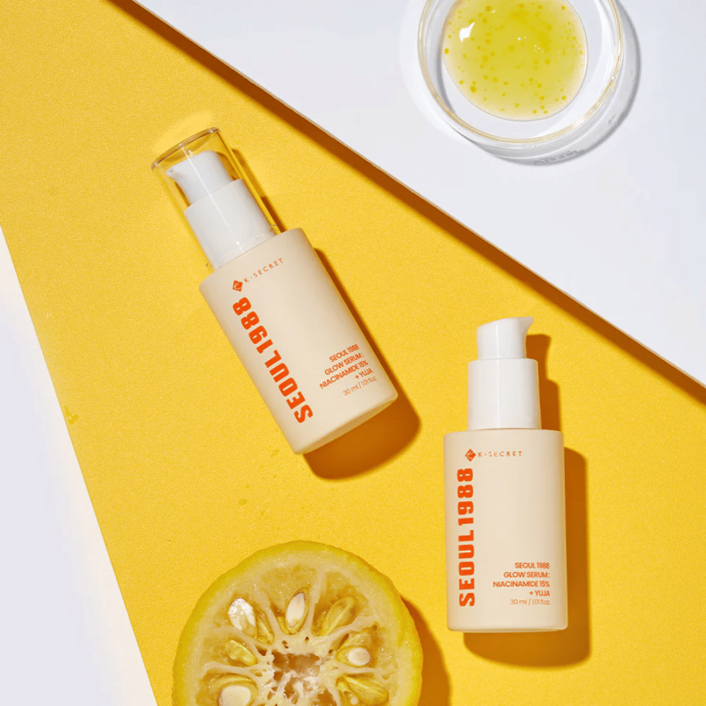 Δύο μπουκάλια Seoul 1988 Glow Serum τοποθετημένα σε κίτρινο φόντο μαζί με ένα μπολ με κίτρινο ορό και ένα μισό εσπεριδοειδές.
