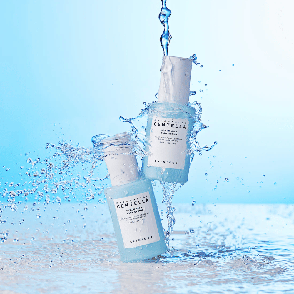 Δύο μπουκάλια SKIN1004 Madagascar Centella Hyalu-Cica Blue Serum χτυπιούνται από έναν καταρράκτη νερού σε ανοιχτόχρωμο μπλε φόντο.