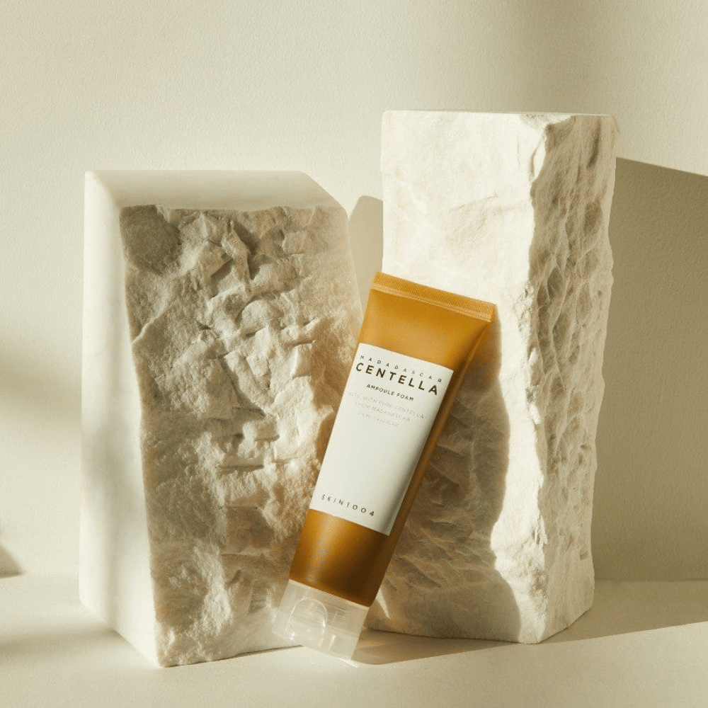 SKIN1004 Madagascar Centella Ampoule Foam στέκεται ενάντια σε φωτεινά πέτρινα μπλοκ σε φυσικό ηλιακό φως, με το καφέ και λευκό σχεδιασμό του σωλήνα και κομψή μινιμαλιστική αισθητική.