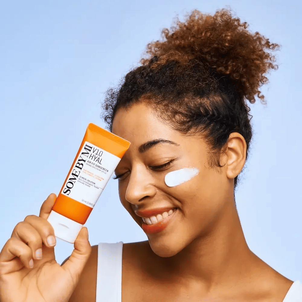 Μια γυναίκα κρατάει έναν σωλήνα με SOME BY MI V10 Hyal Air Fit Sunscreen και έχει μια λευκή κηλίδα αντηλιακής κρέμας στο μάγουλό της. Χαμογελάει χαρούμενα και δείχνει καθαρά το προϊόν, τονίζοντας τη χρήση του και τα οφέλη του για την περιποίηση του δέρματος και την προστασία από τον ήλιο.