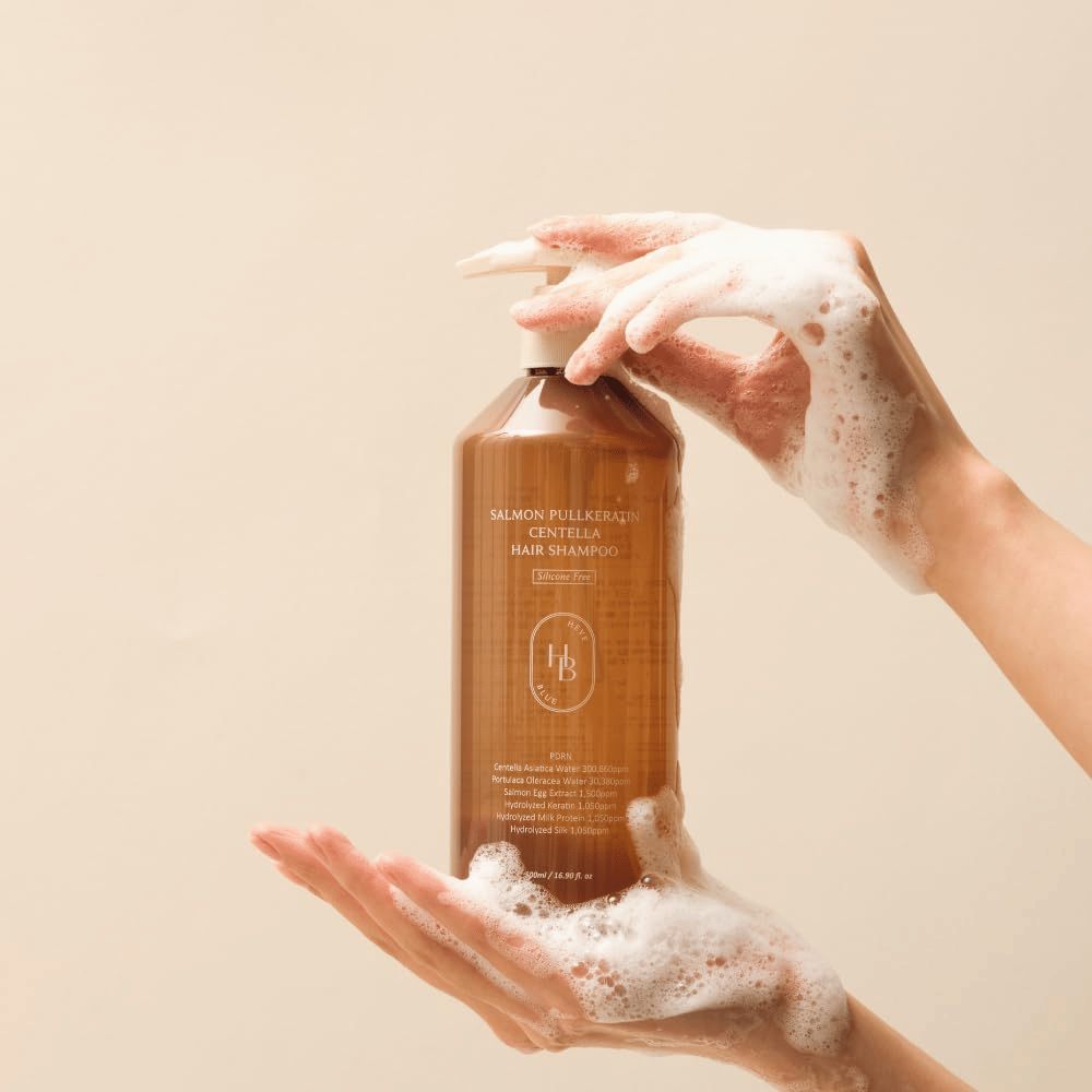 Heveblue Salmon Pullkeratin Centella Hair Shampoo σε καφέ μπουκάλι περιτριγυρισμένο από αφρό. Το προϊόν κρατιέται ανάμεσα σε δύο χέρια και αναδεικνύει μια εμπειρία περιποίησης μαλλιών καθαρισμού και φροντίδας με πλούσιο αφρό.