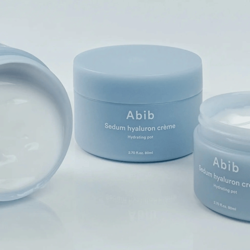Τρία δοχεία του Abib Sedum Hyaluron Crème Hydrating Pot 80 ml σε ανοιχτό μπλε μινιμαλιστικό σχεδιασμό, εκ των οποίων ένα ανοιχτό δοχείο δείχνει την κρεμώδη λευκή υφή.
