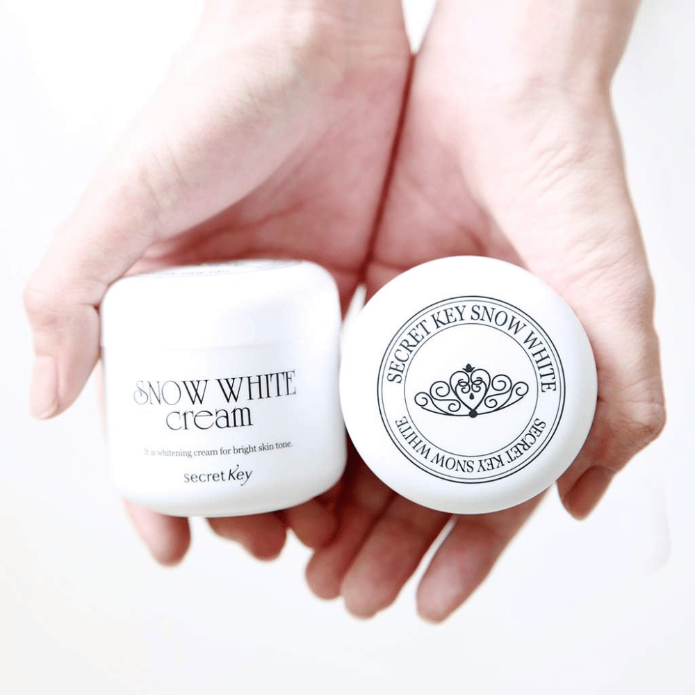 Δύο δοχεία Secret Key Snow White Cream κρατιούνται στα χέρια, το ένα δείχνει το καπάκι και το άλλο την μπροστινή πλευρά.