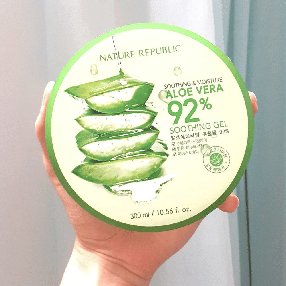 Ένα χέρι κρατάει ένα στρογγυλό δοχείο με Nature Republic Aloe Vera 92% Soothing Gel. Το δοχείο είναι πράσινο με μια εικόνα αλόης και κείμενο που περιγράφει τις καταπραϋντικές και ενυδατικές του ιδιότητες. Το περιεχόμενο είναι 300 ml.