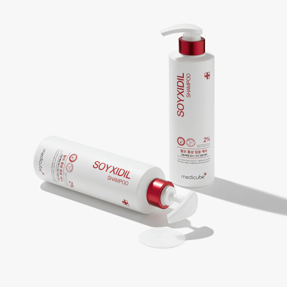 Δύο μπουκάλια του Medicube Soyxidil Shampoo σε λευκό και κόκκινο σχέδιο, το ένα όρθιο και το άλλο ξαπλωμένο με χυμένο σαμπουάν δίπλα.
