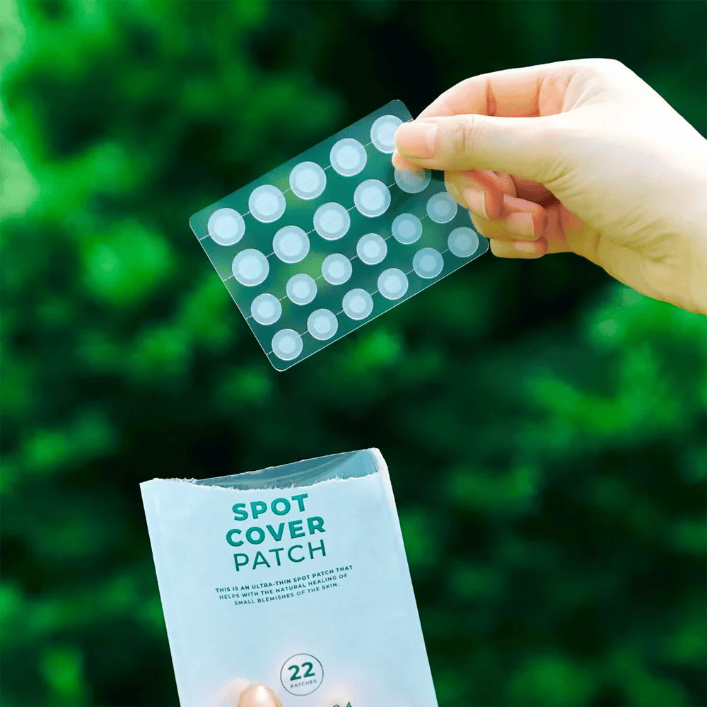 Ένα χέρι κρατάει ένα φύλλο με τα Spot Cover Patches, που είναι διαφανή και στρογγυλεμένα. Στο φόντο φαίνεται ένα θολό πράσινο περιβάλλον. Η συσκευασία δείχνει ένα μέρος της συσκευασίας με το κείμενο "SPOT COVER PATCH" και πληροφορίες ότι το προϊόν περιέχει 22 επιθέματα που βοηθούν στην επούλωση μικρών περιοχών του δέρματος.