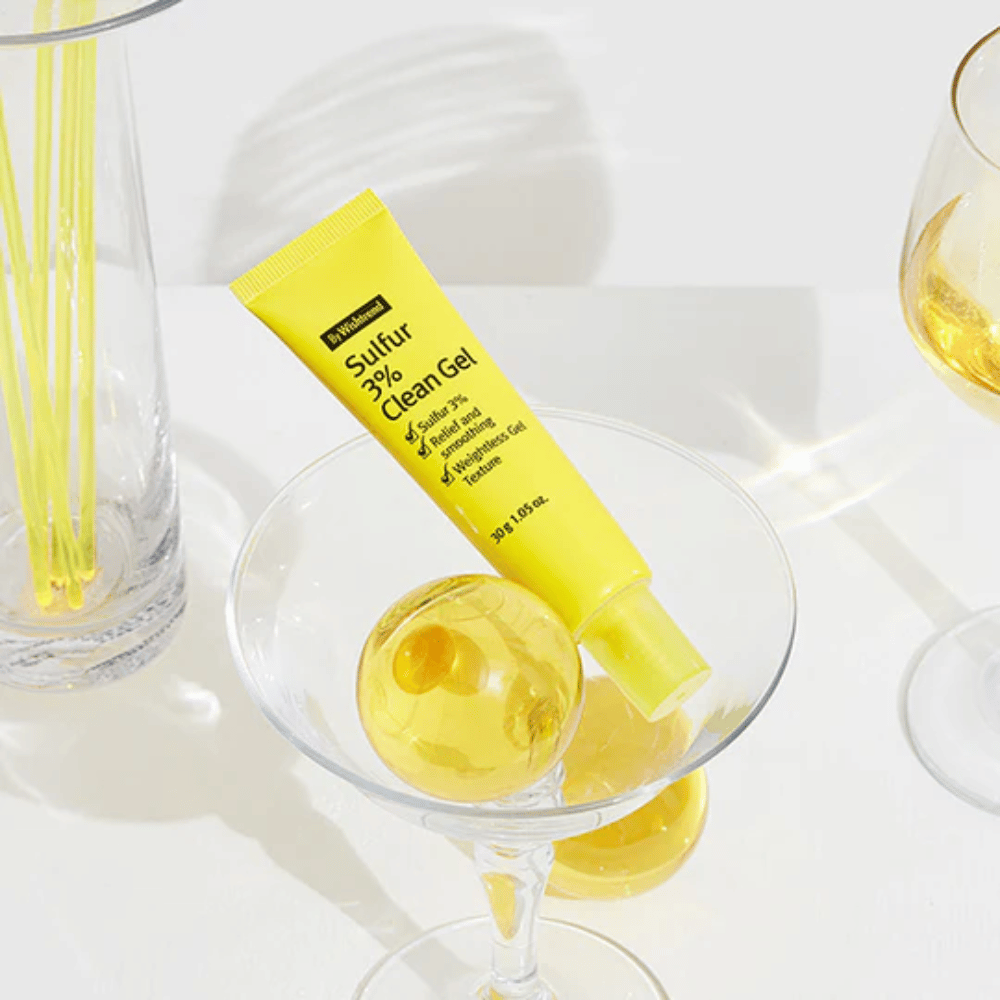 By Wishtrend Sulfur 3% Clean Gel σε κίτρινο σωληνάριο τοποθετημένο σε ποτήρι κοκτέιλ με μια κίτρινη σφαίρα, περιτριγυρισμένο από κομψά ποτήρια σε λευκό φόντο.