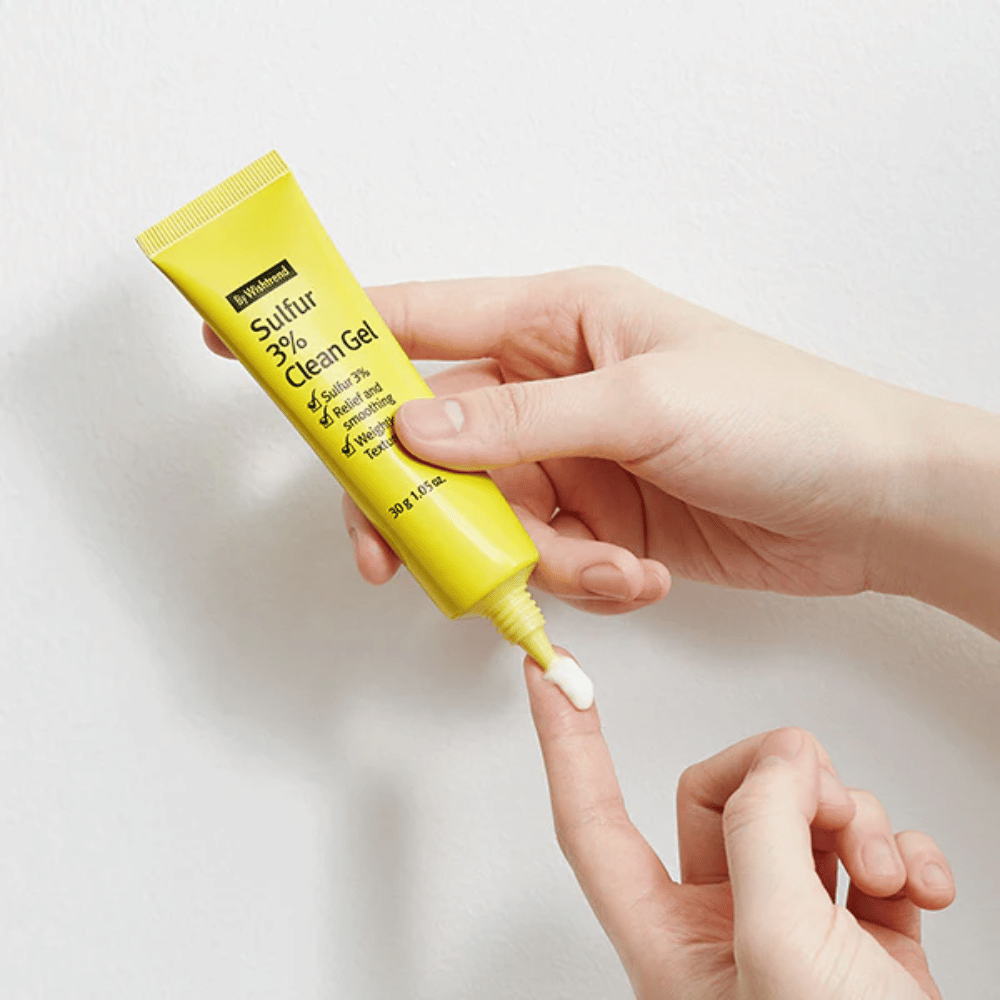 Κοντινή εικόνα χεριών που εφαρμόζουν By Wishtrend Sulfur 3% Clean Gel, ένα κίτρινο σωληνάριο με λευκή γέλη, σε λευκό φόντο.
