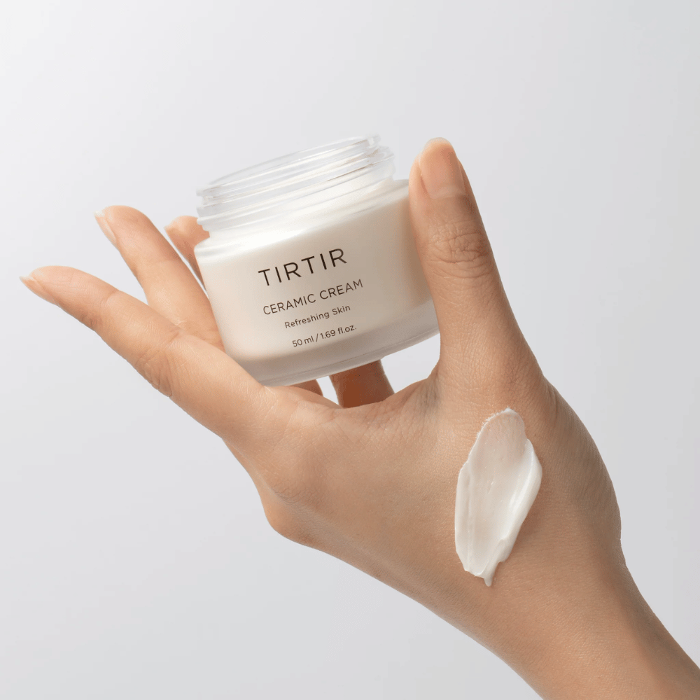 Ένα χέρι κρατάει ένα δοχείο με TIRTIR Ceramic Cream. Στην πίσω πλευρά του χεριού έχει εφαρμοστεί μια ποσότητα από την κρέμα. Η κρέμα φαίνεται να είναι παχιά και ενυδατική. Η συσκευασία αναφέρει ότι το περιεχόμενο είναι για "Refreshing Skin" και η ποσότητα είναι 50 ml / 1.69 fl oz.