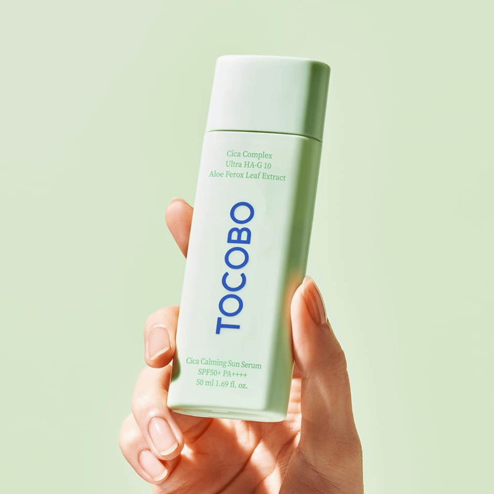 Χέρι κρατάει το TOCOBO Cica Calming Sun Serum με συσκευασία χρώματος μέντας και μπλε κείμενο σε ανοιχτό πράσινο φόντο.