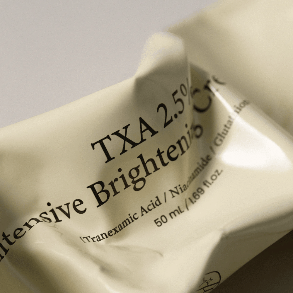 Κοντινό πλάνο του/της AXIS - Y TXA 2.5% Intensive Brightening Cream όπου ο απαλός και μινιμαλιστικός σχεδιασμός του σωληναρίου βρίσκεται στο επίκεντρο. Η εικόνα αναδεικνύει το όνομα του προϊόντος, το Tranexamic Acid και το Niacinamide, καθώς και την κομψή μπεζ συσκευασία.