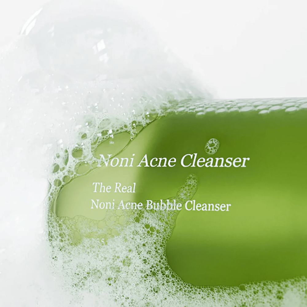 Το μπουκάλι με The Real Noni Acne Bubble Cleanser είναι στο επίκεντρο και περιβάλλεται από άφθονο αφρό. Το φρέσκο πράσινο χρώμα σηματοδοτεί φυσικά συστατικά, ιδανικά για τον καθαρισμό και την καταπολέμηση της ακμής. Ένα ιδανικό προϊόν για καθαρό και διαυγές δέρμα.