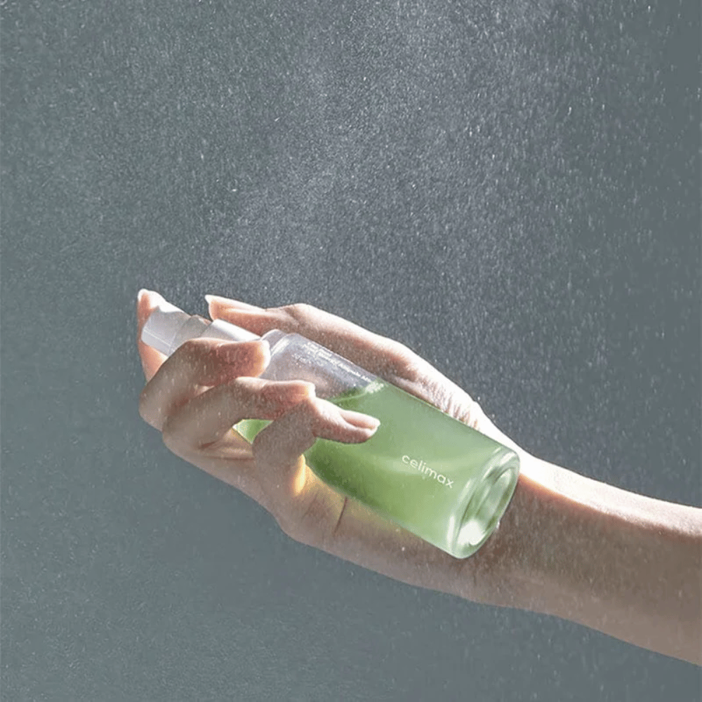 Ένα χέρι κρατάει Celimax Noni Mist μπουκάλι σπρέι και εφαρμόζει το προϊόν σε ένα λεπτό, ομοιόμορφο ψεκασμό στο δέρμα. Το μπουκάλι είναι πράσινο και το προϊόν προωθείται ως "The Real Noni Energy Ampule Mist" για να παρέχει ενέργεια και ενυδάτωση στο δέρμα.