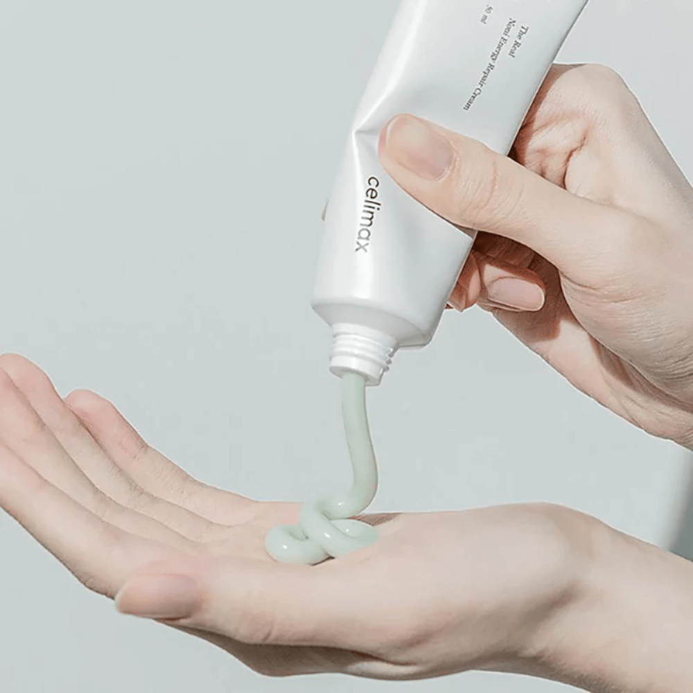 Ένα χέρι κρατάει ένα λευκό σωληνάριο με Celimax Noni Energy Repair Cream, και η κρέμα πιέζεται σε ομοιόμορφη ροή στην παλάμη του χεριού. Η κρέμα είναι ανοιχτοπράσινη και προωθείται για θρέψη και αποκατάσταση του δέρματος.