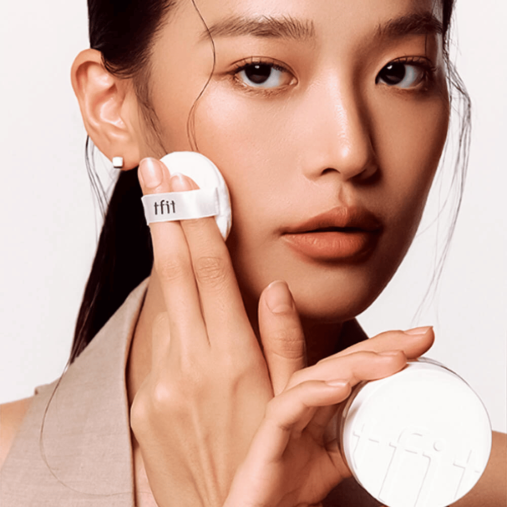 Γυναίκα εφαρμόζει TFIT Translucent Set Finishing Powder #02 Skin Beige με ένα πούφ, κρατώντας ένα λευκό κουτί πούδρας με το λογότυπο TFIT.