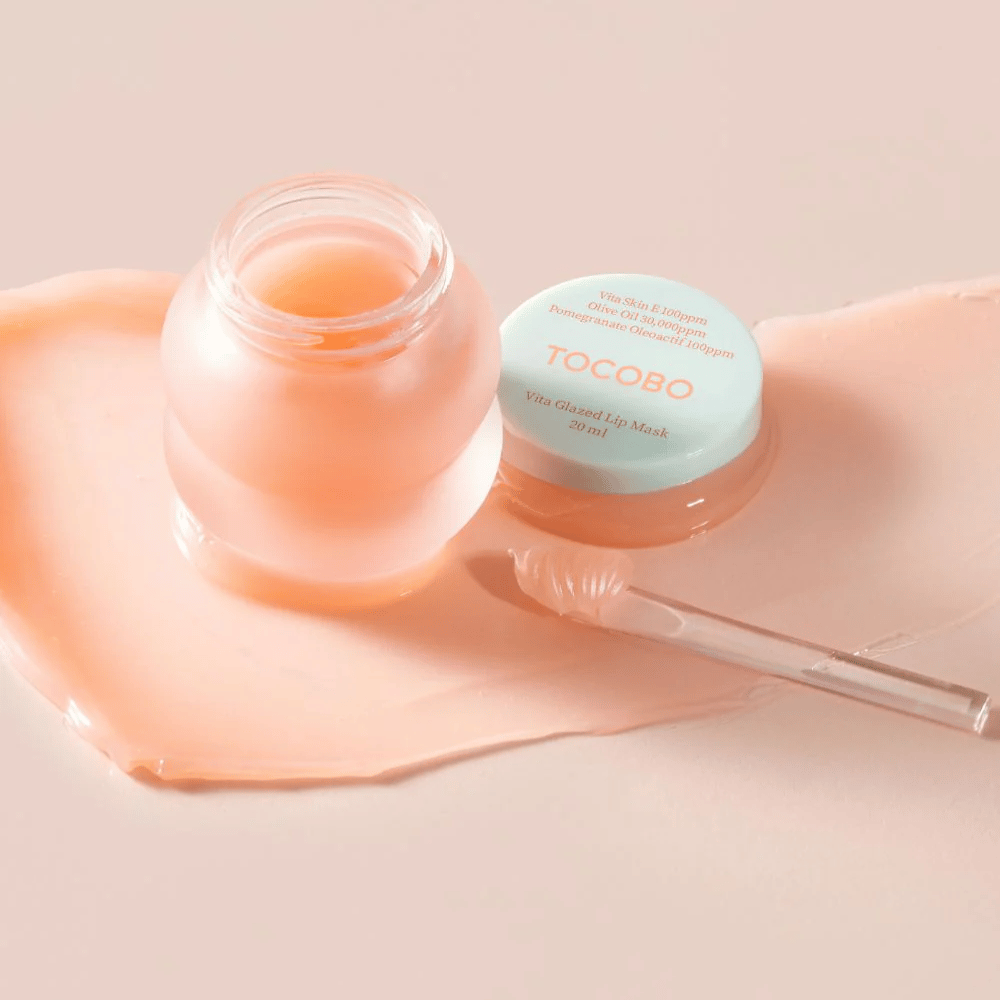 Ένα βάζο με "Vita Glazed Lip Mask" από Tocobo εμφανίζεται. Το βάζο είναι διάφανο με ένα ανοιχτό τιρκουάζ καπάκι και περιέχει μια γυαλιστερή, ροζ μάσκα για τα χείλη. Ένα μικρό σπάτουλα βρίσκεται δίπλα στο βάζο και ένα μέρος της μάσκας είναι απλωμένο στην επιφάνεια. Το προϊόν είναι σχεδιασμένο για να ενυδατώνει και να φροντίζει τα χείλη.