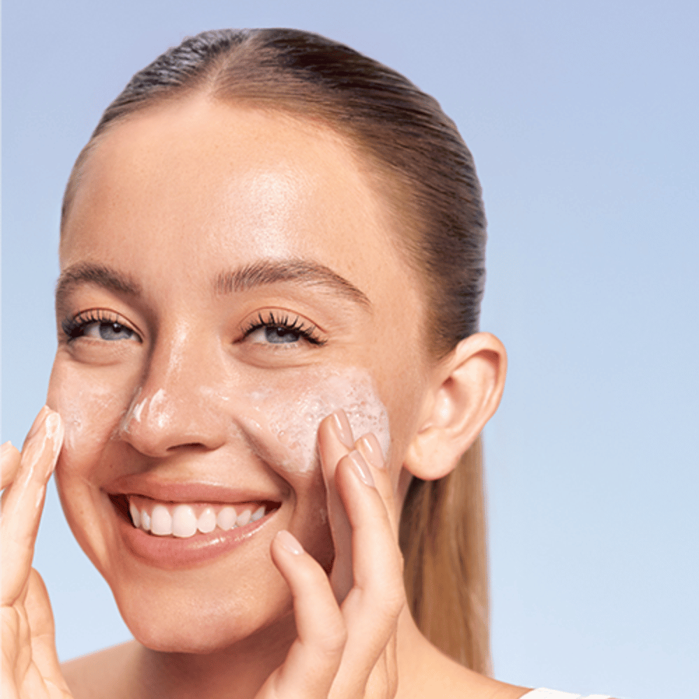 Χαμογελαστή γυναίκα εφαρμόζει LANEIGE Water Bank Gentle Gel Cleanser σε αφρώδη μορφή στα μάγουλα, σε μπλε φόντο.