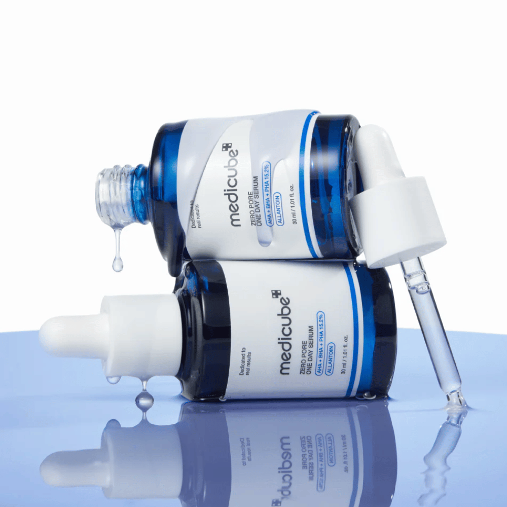 Δύο μπλε μπουκάλια από Medicube Zero Pore One Day Serum που ακουμπούν το ένα πάνω στο άλλο, με πιπέτες και σταγόνες ορού σε μια ανακλαστική μπλε επιφάνεια.