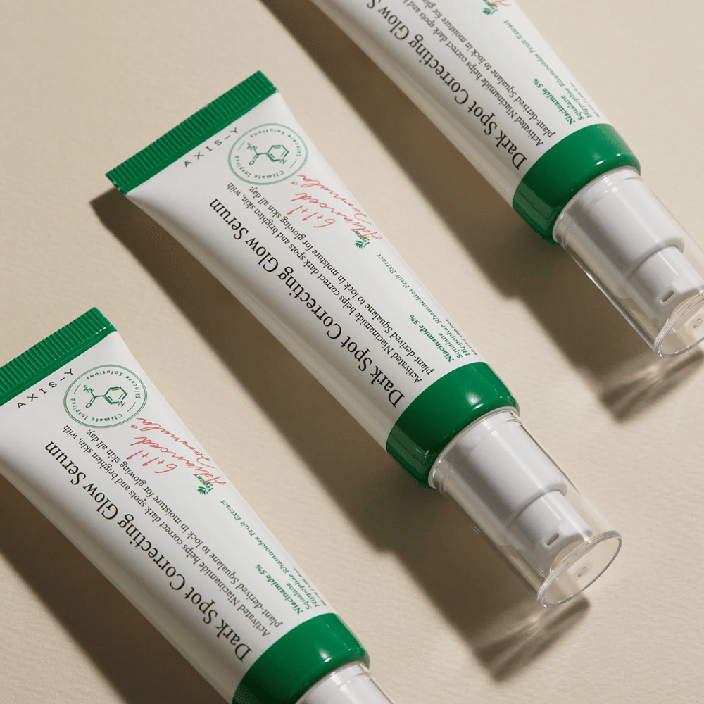 Τρεις σωλήνες από Axis-Y Dark Spot Correcting Glow Serum με πράσινο καπάκι και λευκή συσκευασία βρίσκονται σε ανοιχτόχρωμο υπόβαθρο.