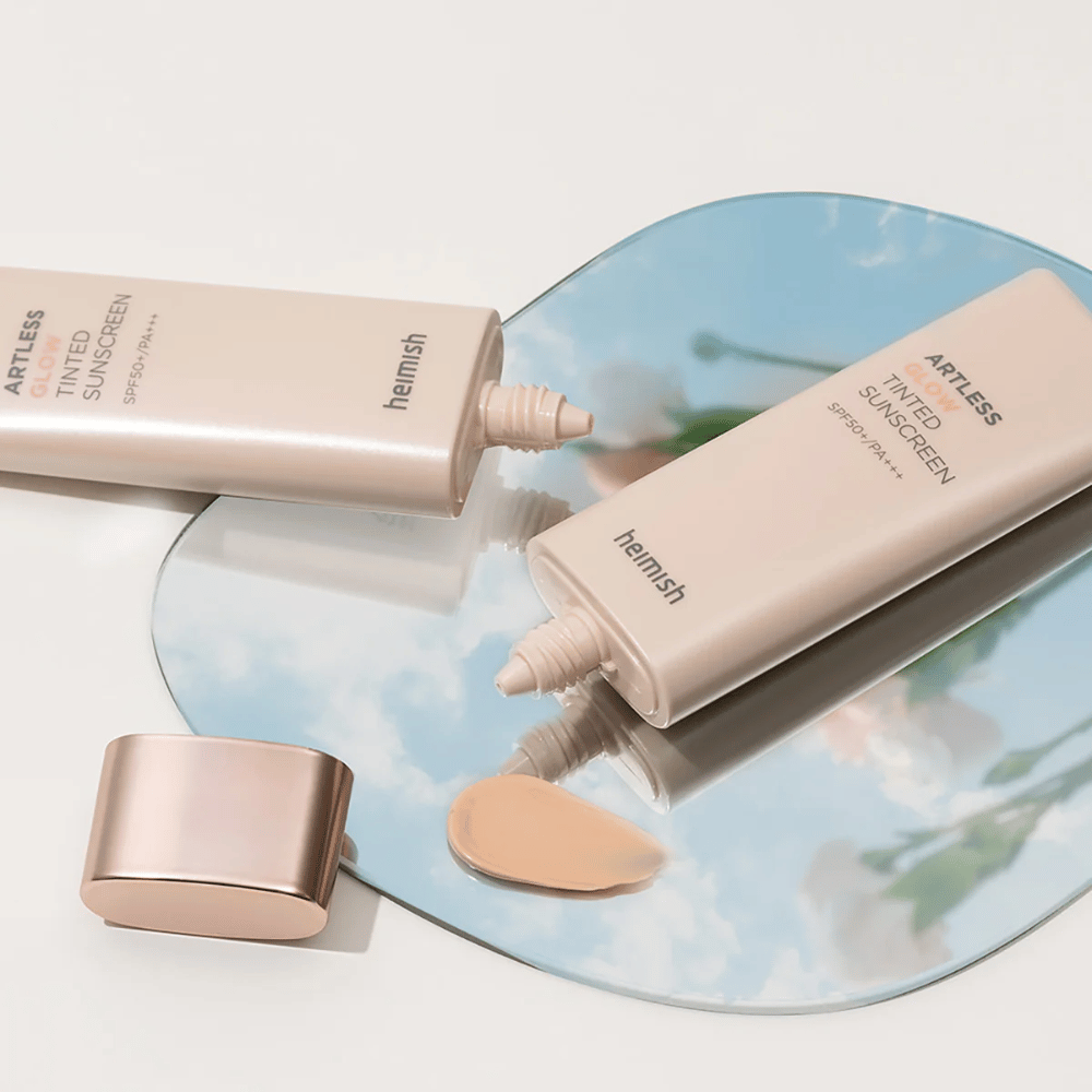 Ένα μπουκάλι heimish Artless Glow Tinted Sunscreen SPF50+ PA+++ 40ml και ένα μπουκάλι concealer πάνω σε έναν καθρέφτη, που δείχνει διαφορετικούς τόνους δέρματος.