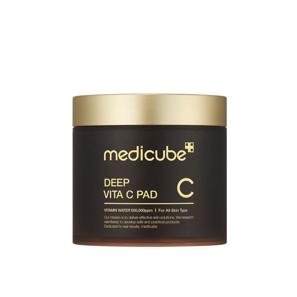 Ένα βάζο από medicube, με την ετικέτα "Deep Vita C Pad", με χρυσό καπάκι και σκούρο σώμα. Το κείμενο στο βάζο αναφέρει βιταμίνη C και καταλληλότητα για όλους τους τύπους δέρματος. Το φόντο είναι ουδέτερο.