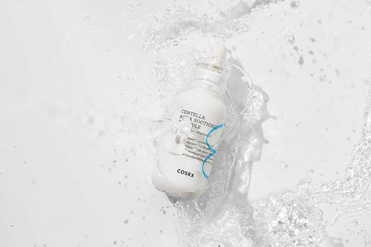 Ένα μπουκάλι COSRX Centella Aqua Soothing Ampoule 40ml που πιτσιλά σε μια λευκή επιφάνεια, μειώνει την ερυθρότητα και προσφέρει καταπραϋντική ενυδάτωση.