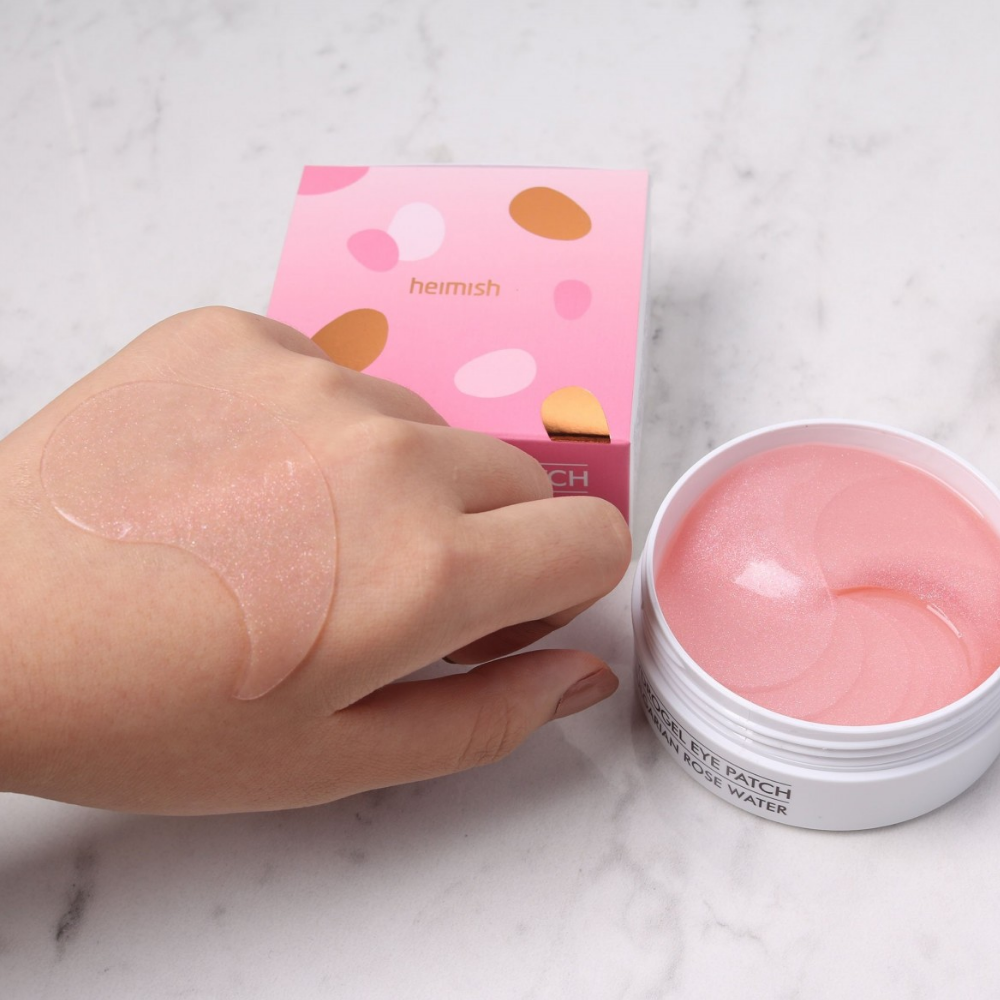 Ένα χέρι που κρατάει μια ροζ Bulgarian Rose Hydrogel Eye Patch 60st από heimish και εφαρμόζει μάσκα ματιών.