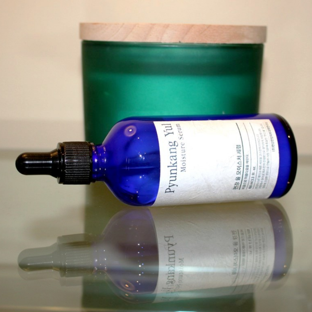 Ένα μπουκάλι Pyunkang Yul Moisture Serum 100ml εξισορροπεί τα επίπεδα υγρασίας σε ένα τραπέζι δίπλα σε ένα κερί.